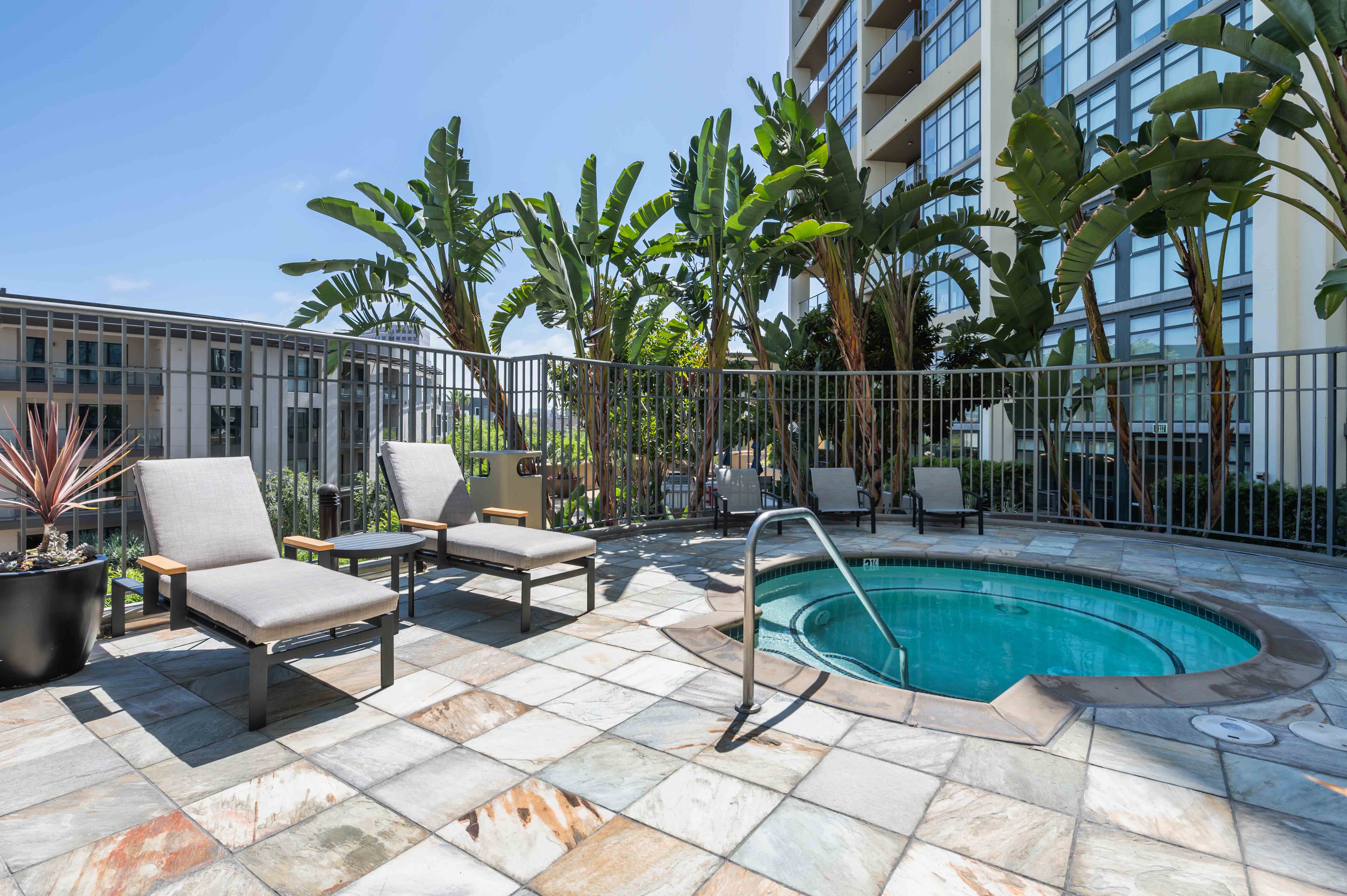 Pet-Friendly Apartments in Irvine, CA - Astoria at Central Park West - Spa with Lounge Seating, Surrounded by Grey Fencing, Potted Plants, and Tropical Trees.